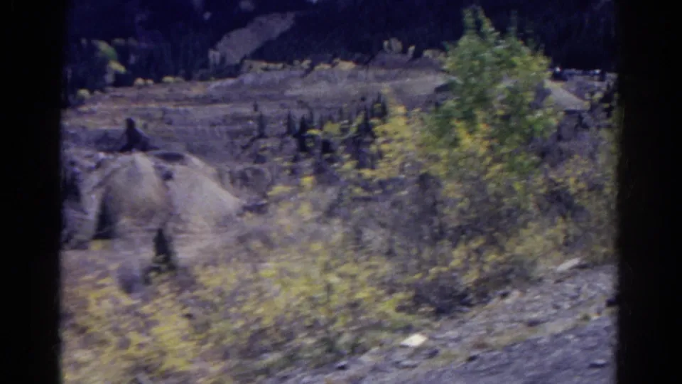 1967 Colorado 8mm frame looking across rocky quarry terrain, scattered conifers, a dirt track, and excavated earth in a mountain valley.