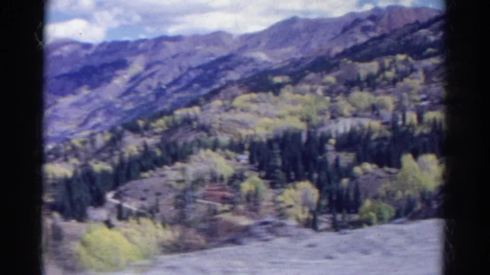 1967 Colorado 8mm frame showing a wider mountain valley with evergreen clusters, pale aspens, rocky ridges, and open sky.