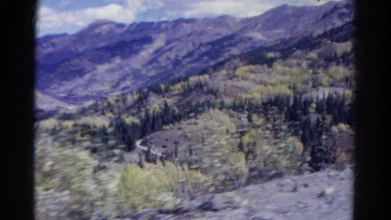 1967 Colorado 8mm frame of a mountain valley seen from a moving car, with a curving road, evergreens, aspens, and distant ridges.