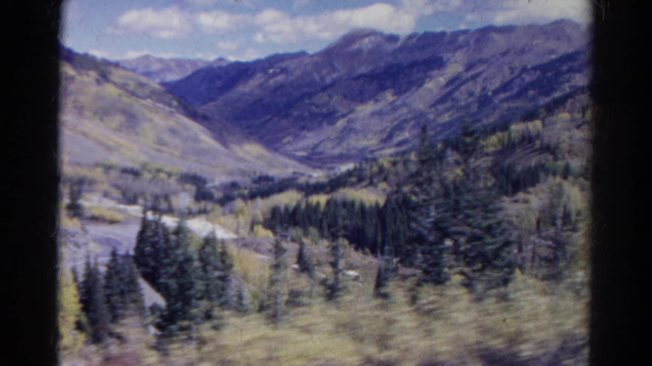 Broad mountain valley with forest patches, pale aspens, road, and distant ridges.