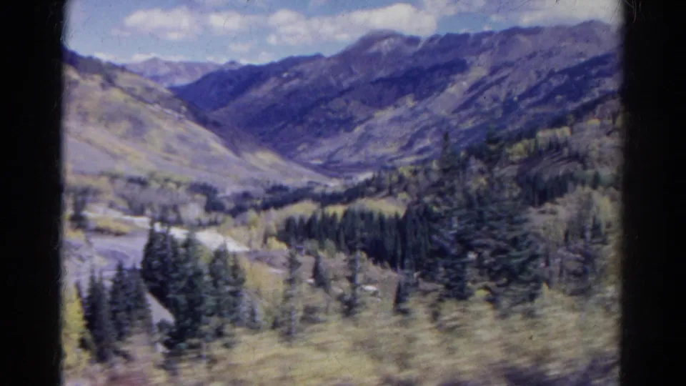 1967 Colorado home-movie view across a high mountain valley, with forested slopes, pale aspen stands, and a road below.