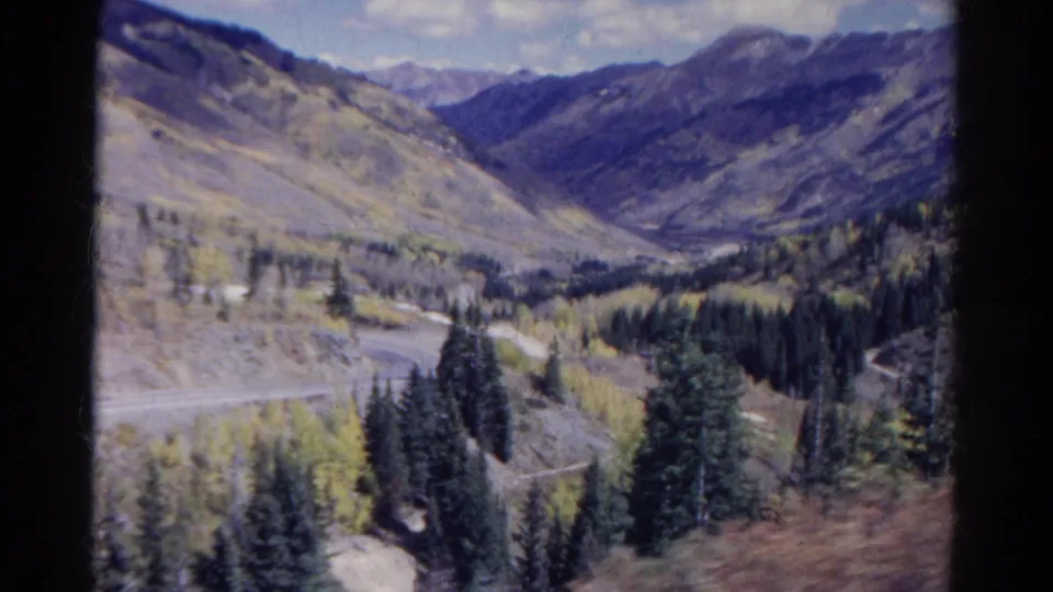 Moving-car 8mm view in Colorado, showing a steep mountainside, roadside curve, dark evergreens, aspens, and a distant valley.