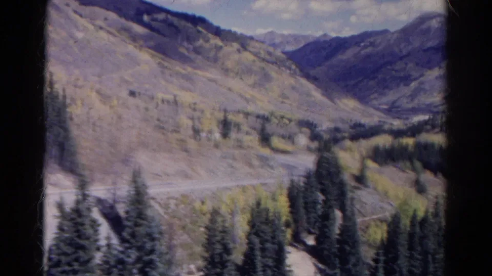 1967 Colorado 8mm frame from a moving car, with motion-blurred trees, a roadside bend, open mountain slopes, and distant ridges.