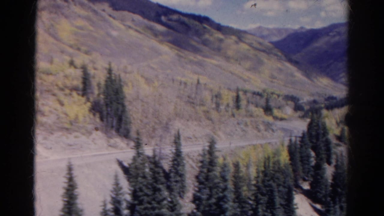 1967 Colorado Super 8 frame showing a dirt road along a mountain slope with evergreens, pale autumn foliage, and distant ridges.