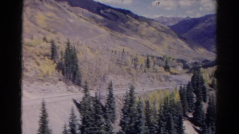 1967 Colorado Super 8 frame showing a dirt road along a mountain slope with evergreens, pale autumn foliage, and distant ridges.
