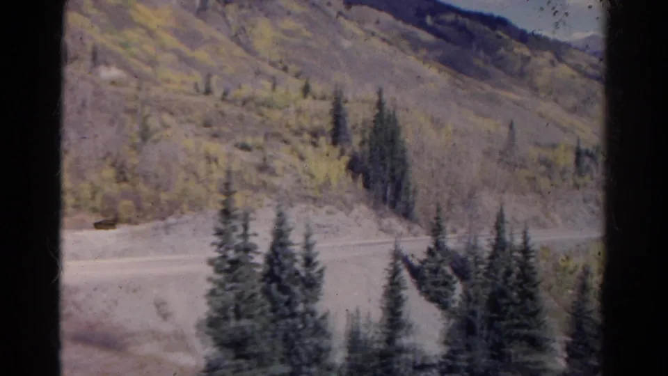 1967 Colorado mountain-road view with evergreens, muted autumn foliage, and a small dark shape near the roadside that is not clearly identifiable.
