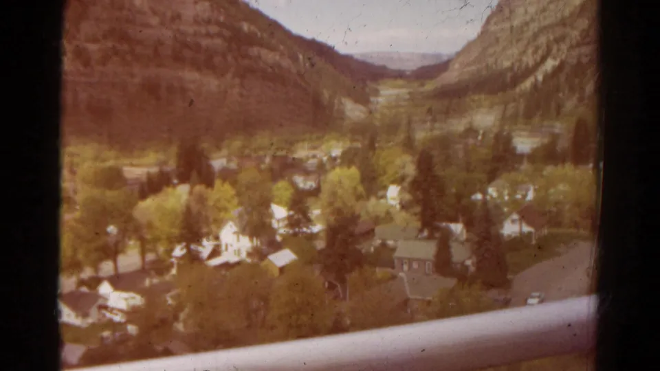 1967 Colorado 8mm view of a valley settlement with houses, trees, steep hills, and a railing in the foreground.