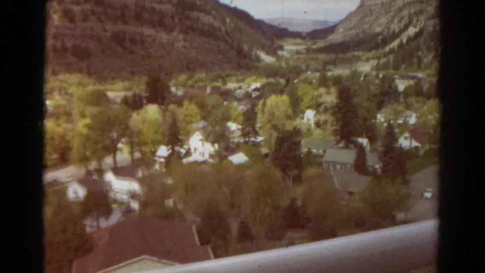 1967 Colorado home-movie frame of a small valley town with homes, trees, roads, surrounding hills, and a railing below.