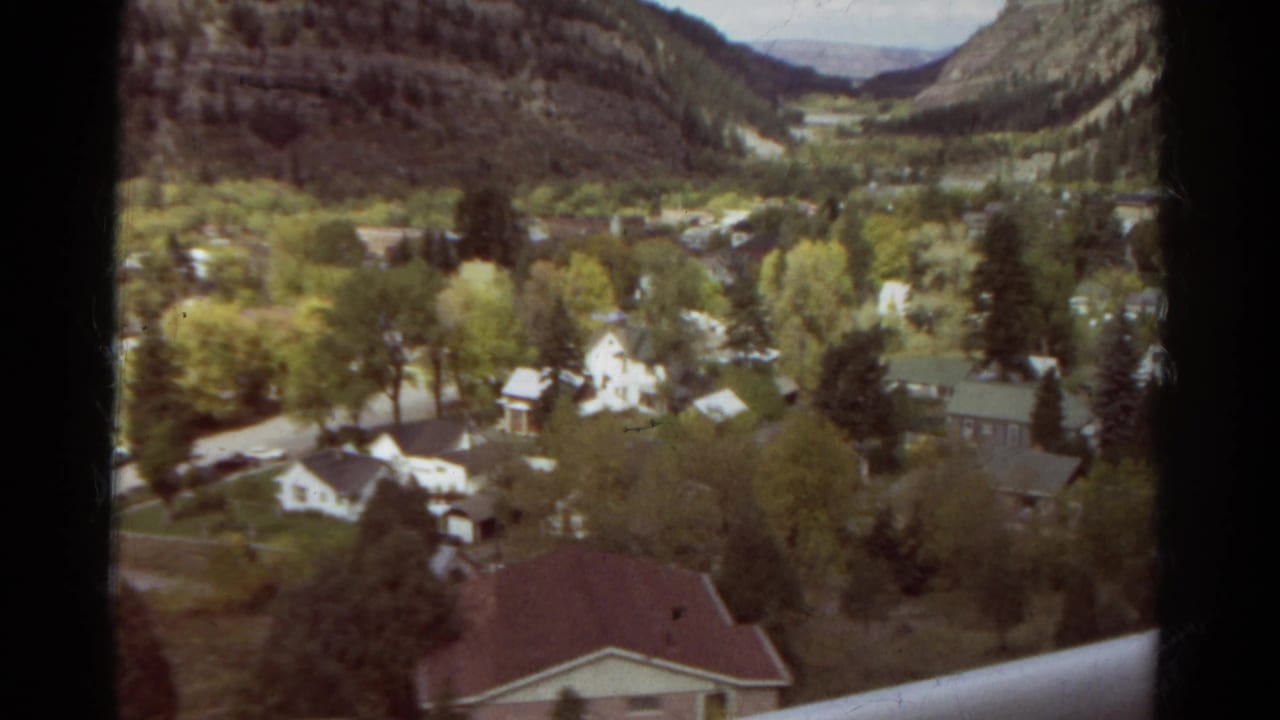 1967 Colorado 8mm view looking down over a mountain-town neighborhood with houses, green trees, roads, and hills beyond.