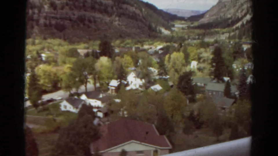 1967 Colorado 8mm view looking down over a mountain-town neighborhood with houses, green trees, roads, and hills beyond.