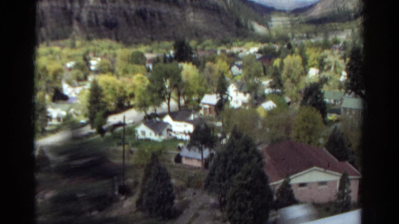 Elevated 1967 Colorado view of tree-lined residential streets, small houses, a red-roofed foreground building, and mountain slopes.