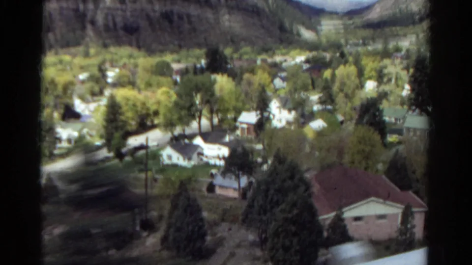 Elevated 1967 Colorado view of tree-lined residential streets, small houses, a red-roofed foreground building, and mountain slopes.