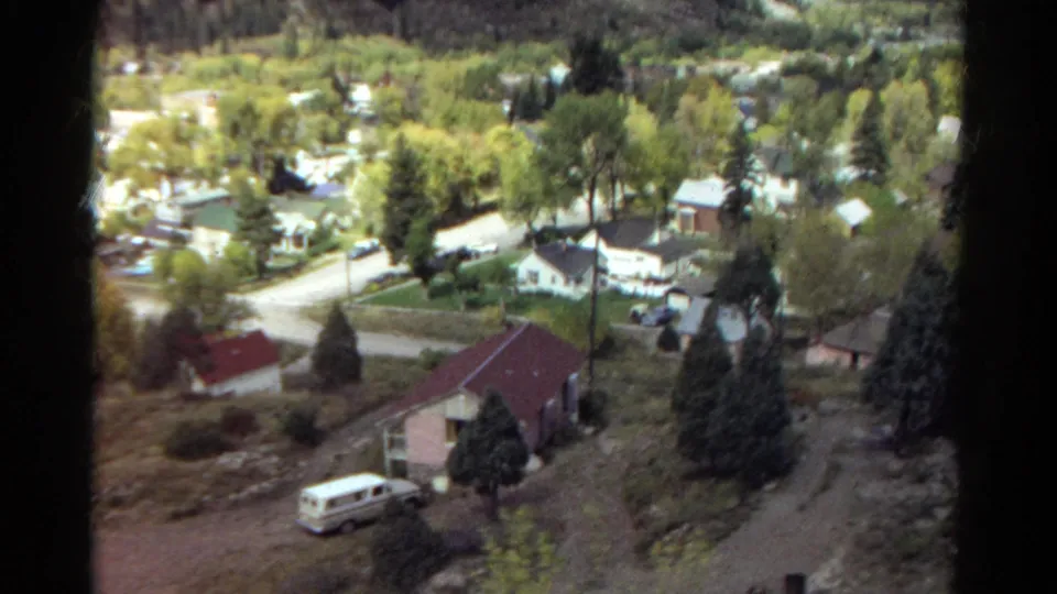 1967 Colorado residential view with a red-roofed house, curved driveway, evergreen trees, and a light station wagon parked below.