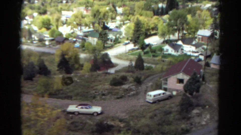 Elevated 1967 Colorado view of classic cars near a residential house, with streets, trees, and valley homes in the background.