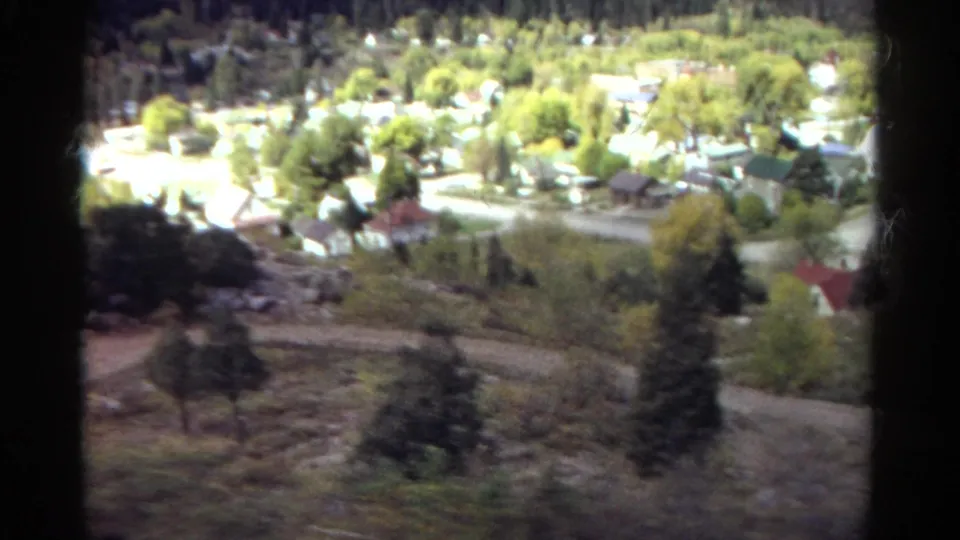 Elevated 1967 Colorado home-movie frame showing a hillside road, clustered houses, trees, and forested slopes beyond.