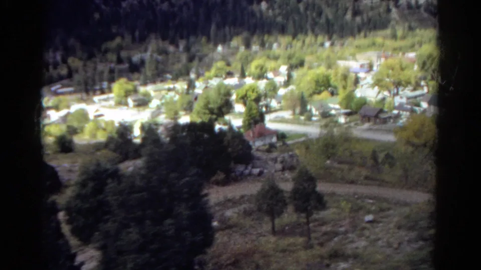 1967 Colorado valley view with a small neighborhood, pale roads, roofs, trees, and shadowed hillside foreground in soft 8mm color.