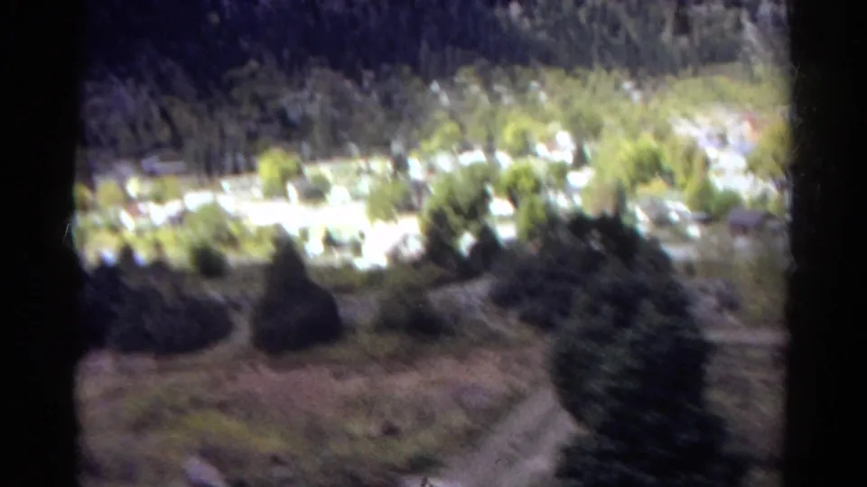 Soft, motion-blurred 1967 Colorado home-movie frame with houses, trees, hillside terrain, and a pale road at the edge of town.