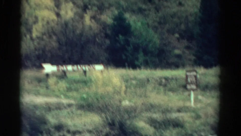 Soft 8mm frame of the Box Canyon sign across a grassy Colorado clearing, with brush and trees behind it.