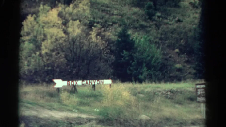 1967 Colorado 8mm frame showing a white Box Canyon arrow sign along a grassy roadside with dense trees behind it.