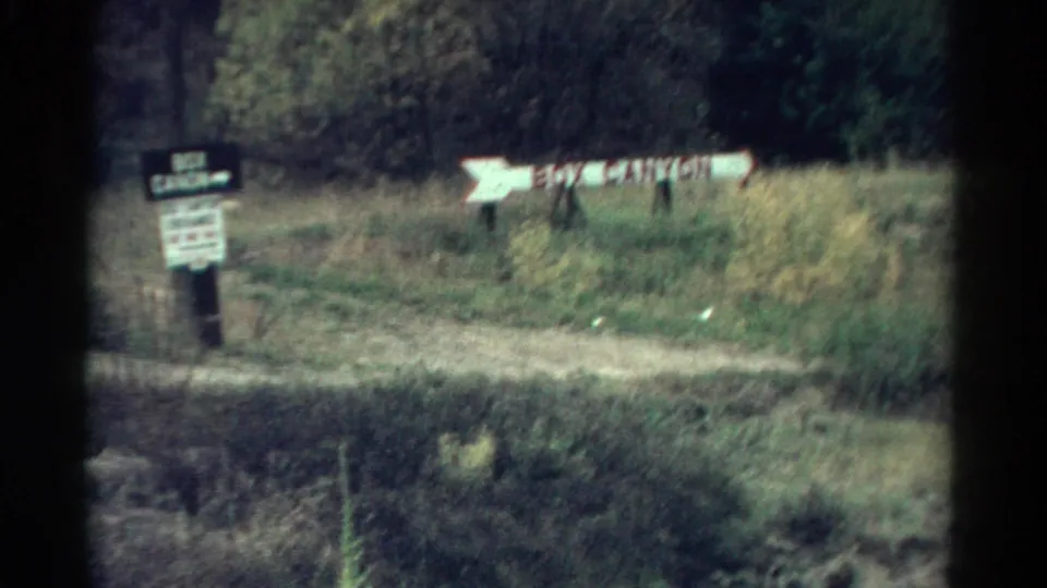 Home-movie frame showing Box Canyon directional signage beside a path, with grass, trees, and dark film edges visible.