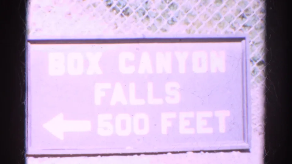 Close 1967 Colorado frame of a Box Canyon Falls 500 Feet directional sign with a left arrow beside chain-link fencing.