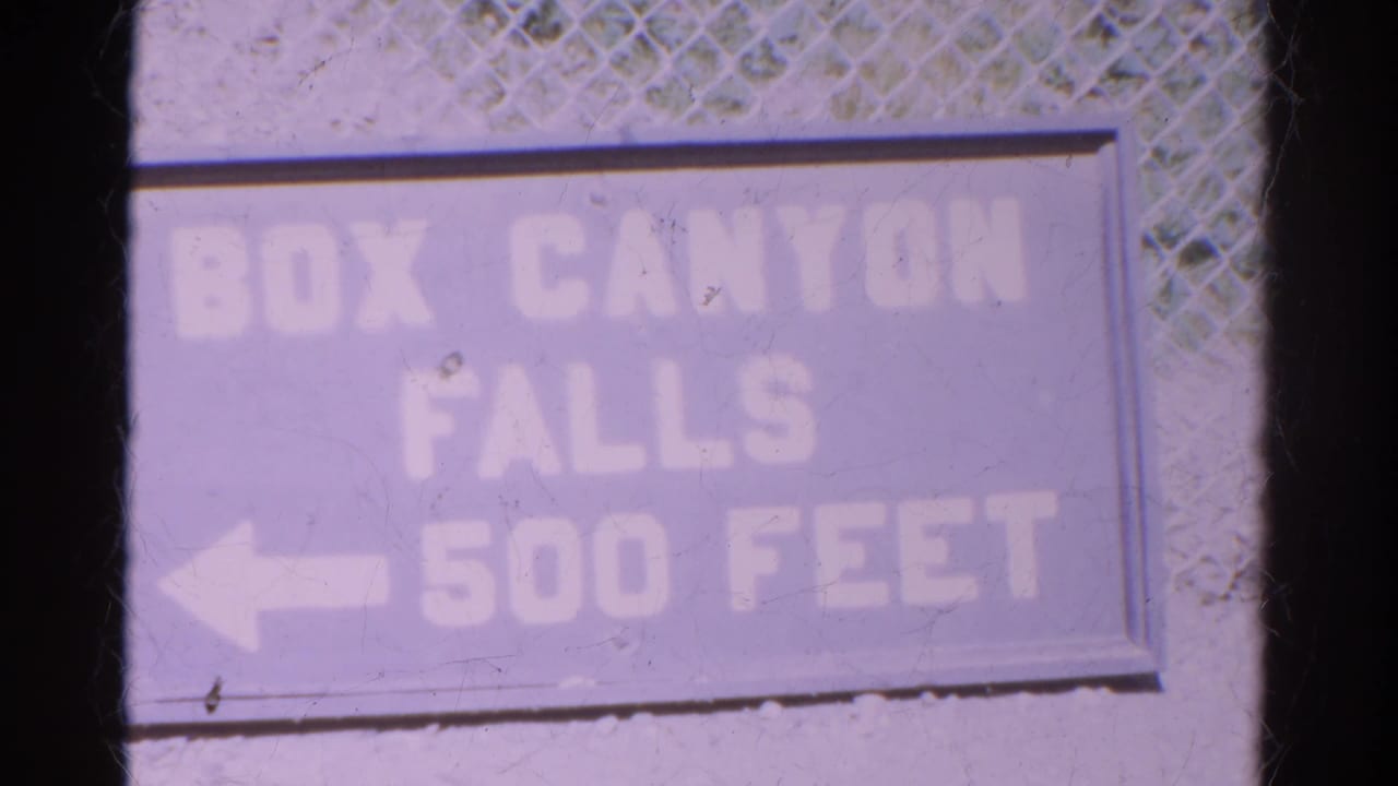 Faded directional sign for Box Canyon Falls, 500 feet away, filmed in a soft, purple-shifted 8mm frame.