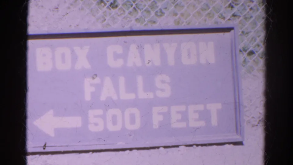 Faded directional sign for Box Canyon Falls, 500 feet away, filmed in a soft, purple-shifted 8mm frame.