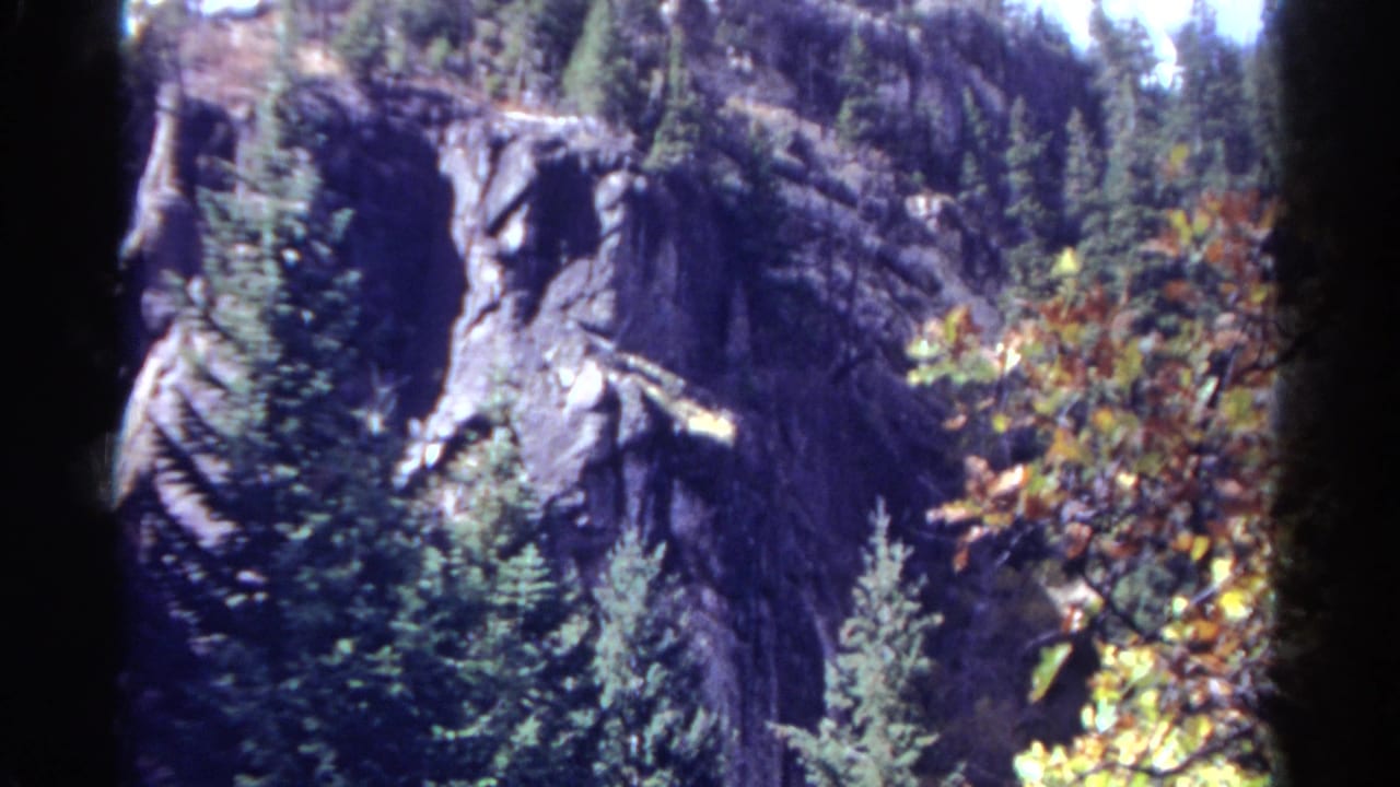 1967 Colorado 8mm view of a steep rock wall with conifer trees below and autumn foliage along the side.