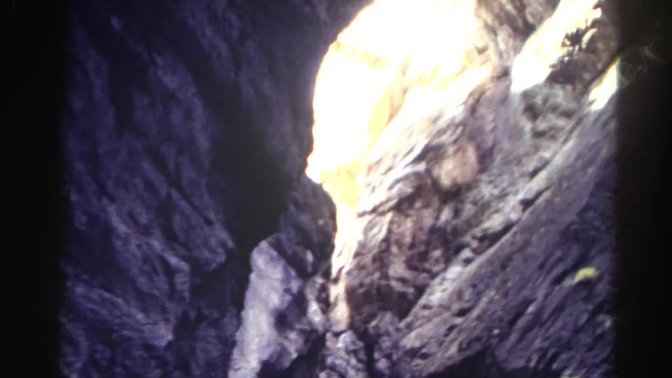 Blue-tinted 8mm view from the canyon bottom toward a bright sunlit opening above rugged Colorado rock.