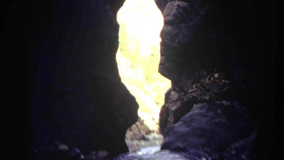 1967 Colorado 8mm frame looking from a dark canyon toward an overexposed sunlit rock gap and water at the base.