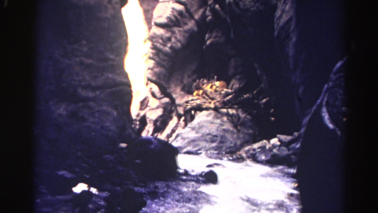 1967 Colorado 8mm frame of a narrow rocky canyon with stream water, boulders, and a sunlit wall opening.