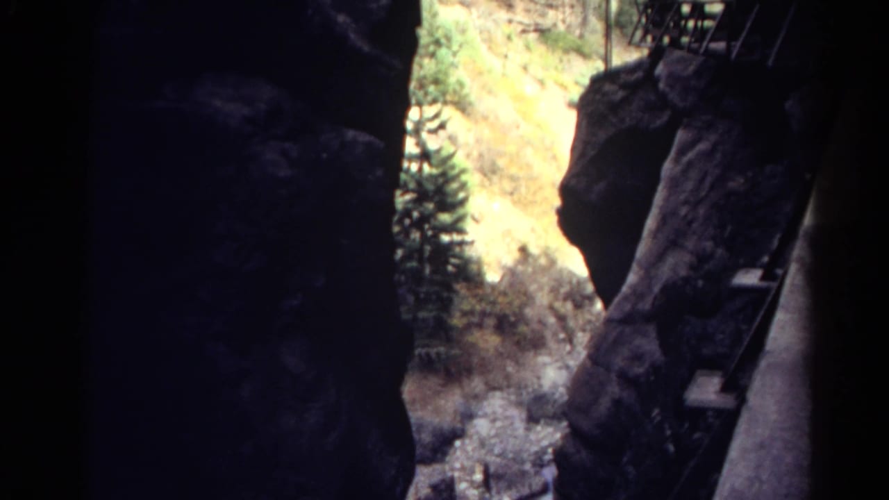 Inside a dark Colorado rock passage, the view shifts toward a bright mountain slope, evergreen trees, and metal climbing infrastructure.