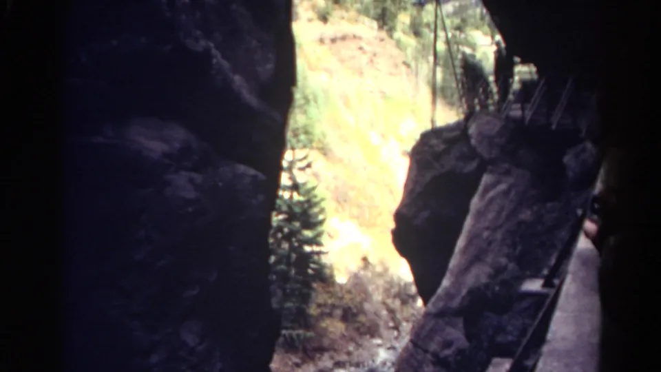 1967 Colorado home-movie frame shows a shadowed cave-like corridor opening onto a rugged slope with trees and ladder sections.