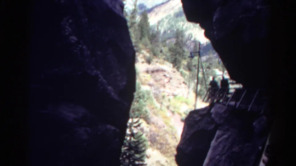 Small hikers are visible on a Colorado rock route with metal ladders or railings, seen from inside a dark cave opening.