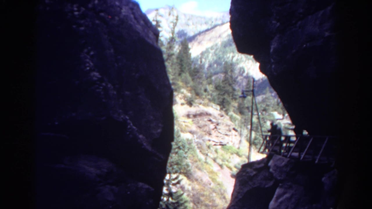 Shadowed canyon opening looking toward bright mountains, trees, and a small overlook platform.