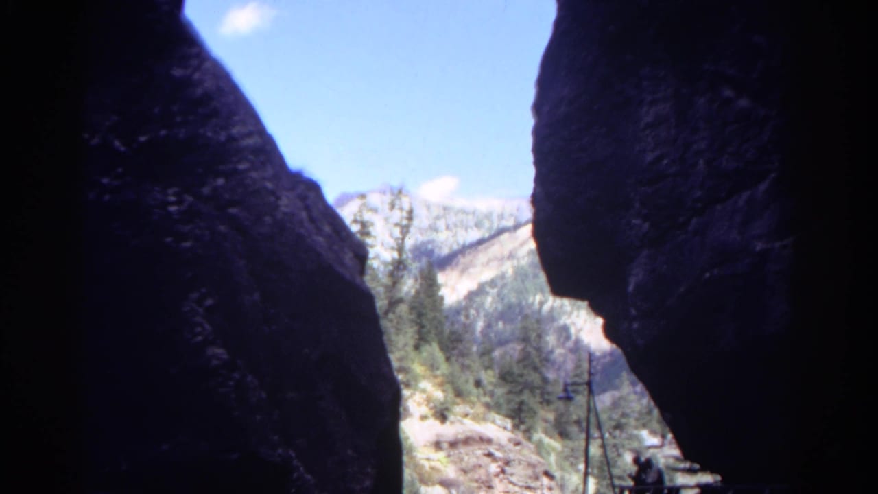 Rock walls frame a sunlit mountain slope with trees, sky, and a small platform at right.