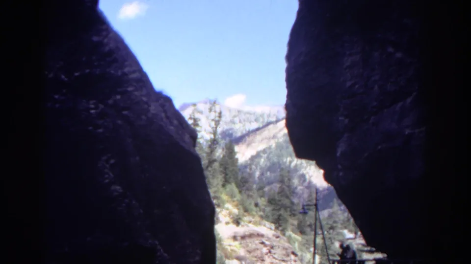 1967 Colorado 8mm view through shadowed canyon walls toward a sunlit mountain slope, trees, blue sky, and distant visitors.