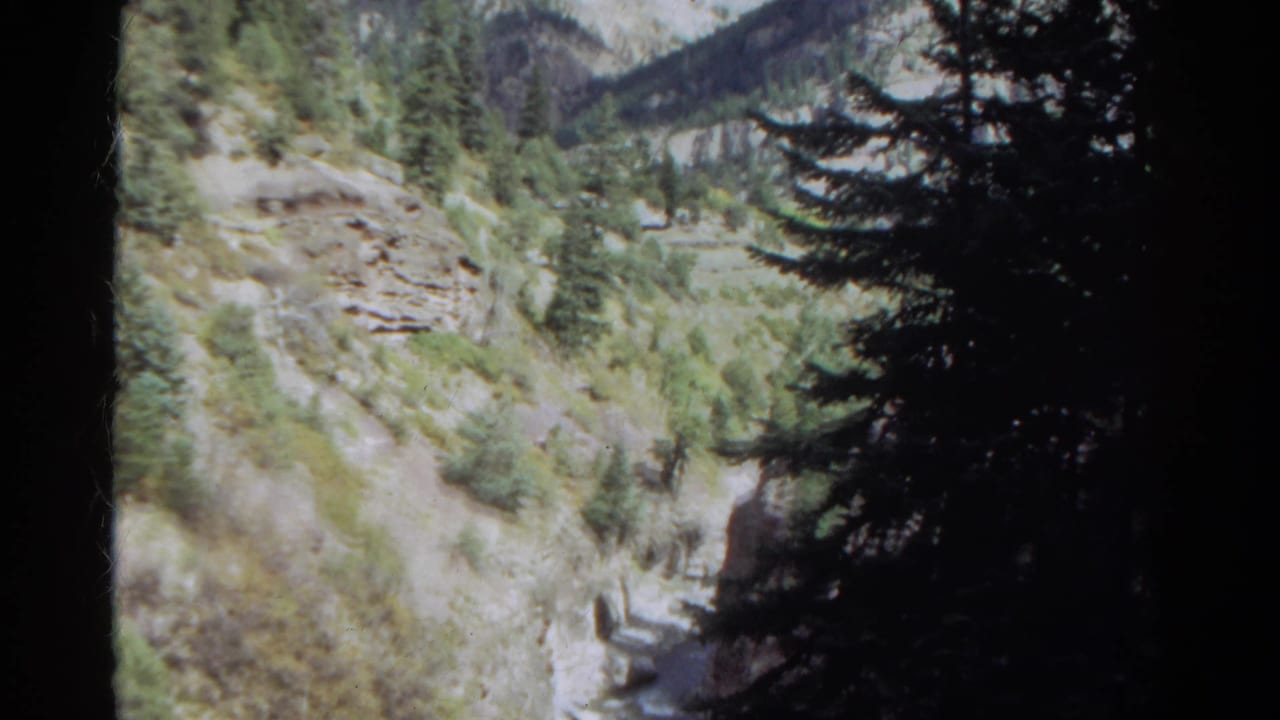 1967 Colorado 8mm frame showing a rocky mountain valley and pine forest from a shaded handheld viewpoint.