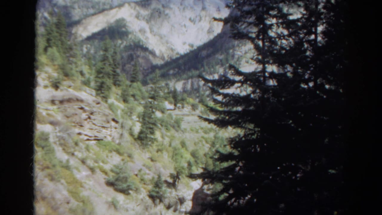 Handheld 8mm view of Colorado pines, a rocky slope, and a narrow ravine or streambed in a mountain valley.