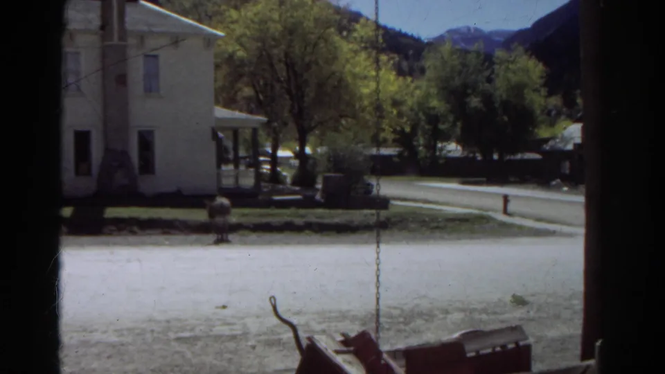 1967 Colorado mountain-town view with a white house, roadside, trees, porch swing foreground, and distant peaks.