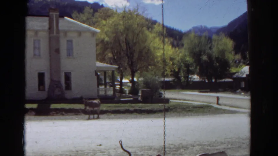 1967 Colorado home-movie still with a horse near a white house and porch, viewed past a dark window or porch edge.