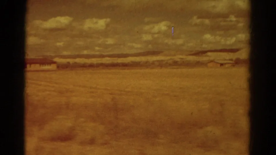 1967 Colorado travel frame showing a broad field, distant hills, cloudy sky, and a small building near the left edge of the frame.