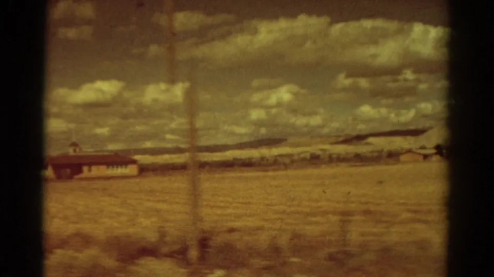Rural Colorado 8mm frame with a small building, open field, distant hills, cloudy sky, and vertical film mark through the scene.