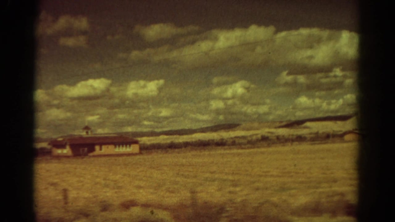 1967 Colorado 8mm frame showing a lone low building beyond a dry field under a broad cloudy sky.