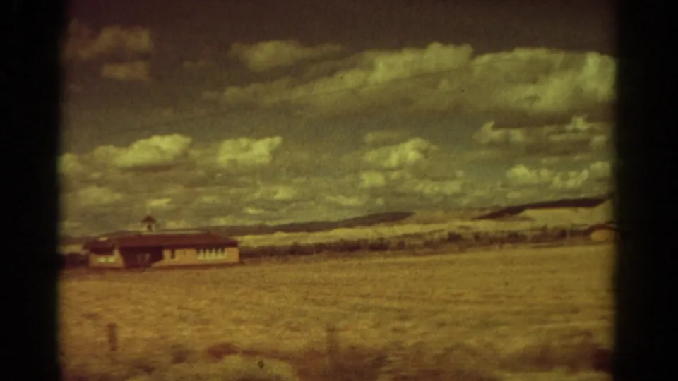 1967 Colorado 8mm frame showing a lone low building beyond a dry field under a broad cloudy sky.