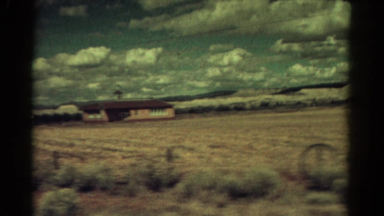 Roadside view of a low building and dry field with clouds and distant hills.