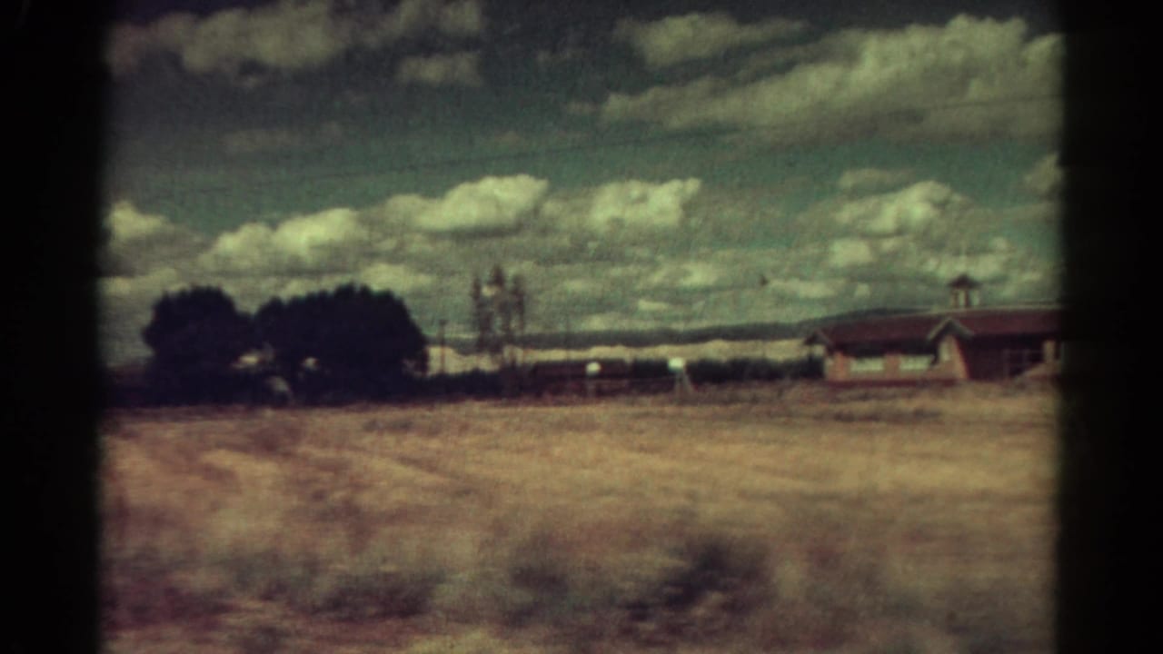 1967 Colorado rural view with a dry open field, dark trees, fence posts, low hills, and part of a house at the right edge.