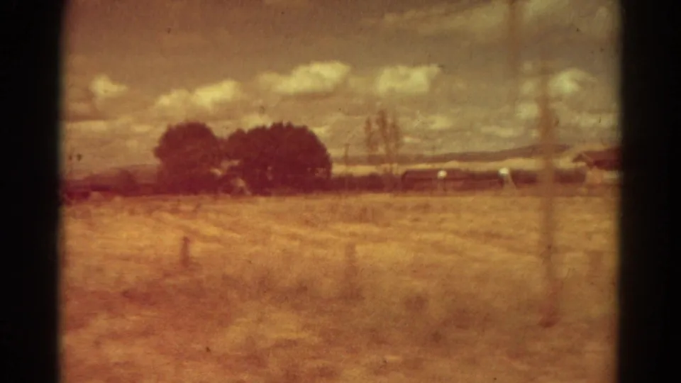 Warm color-shifted 8mm frame of a Colorado field with dark trees, low rural structures, distant hills, and scattered clouds.