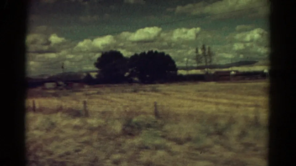 1967 Colorado 8mm view of a dry field with fence posts, dark trees, low buildings, distant hills, and mottled clouds.