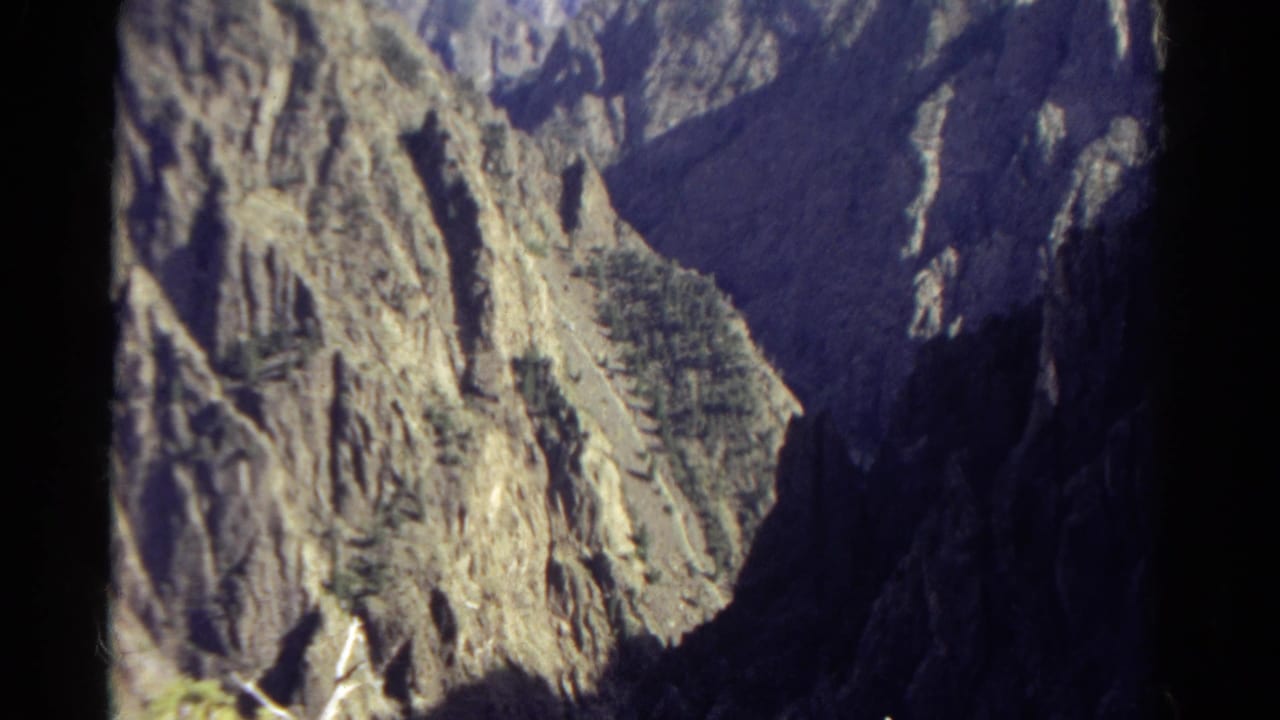 1967 Colorado 8mm frame showing rugged canyon cliffs, evergreen patches, and strong sunlight against dark shadowed rock.